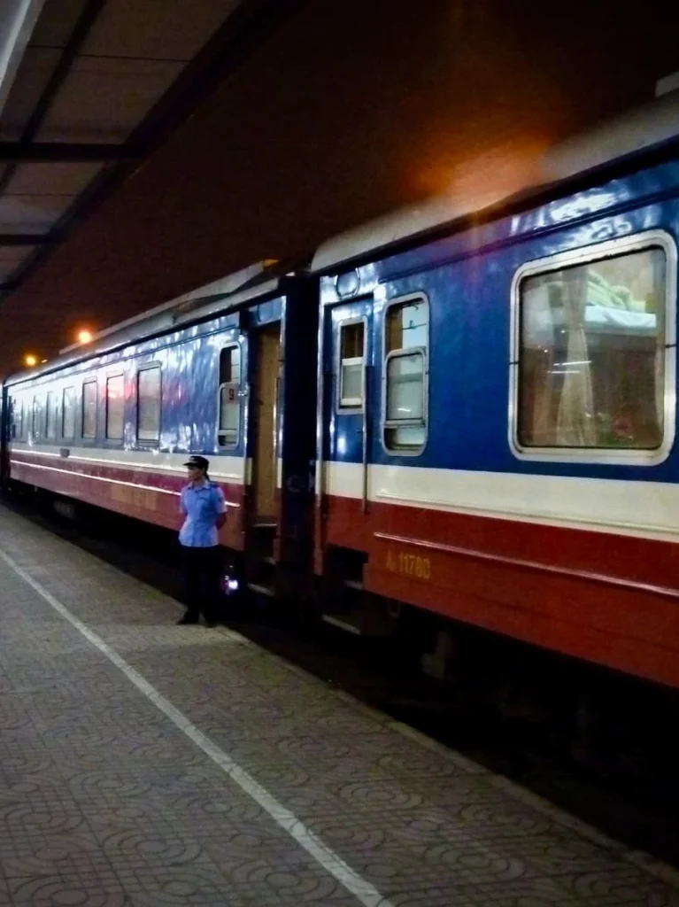 Reunification SE1 train exterior at Hanoi train station with the main train having a blue, white and red stripe on the outside and a female conductor standing outside.