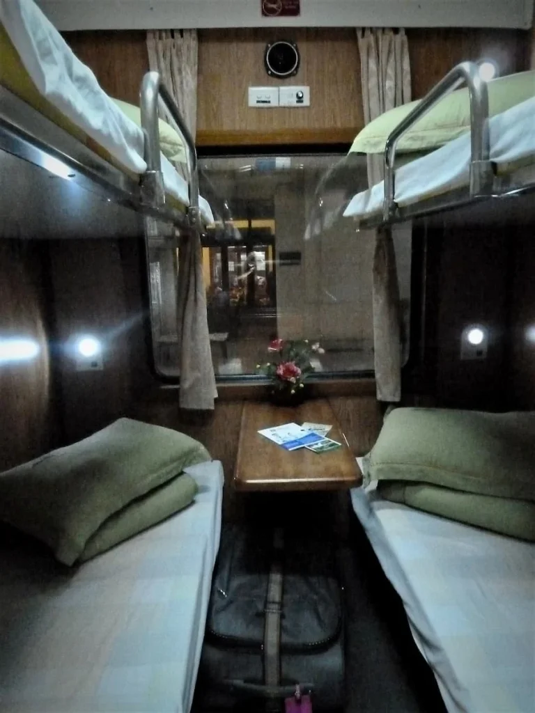 Reunification Train cabin with 4 berths and white sheets and mattresses. the pillows have light green covers and there is a small wooden fold-up table in the middle of the berths