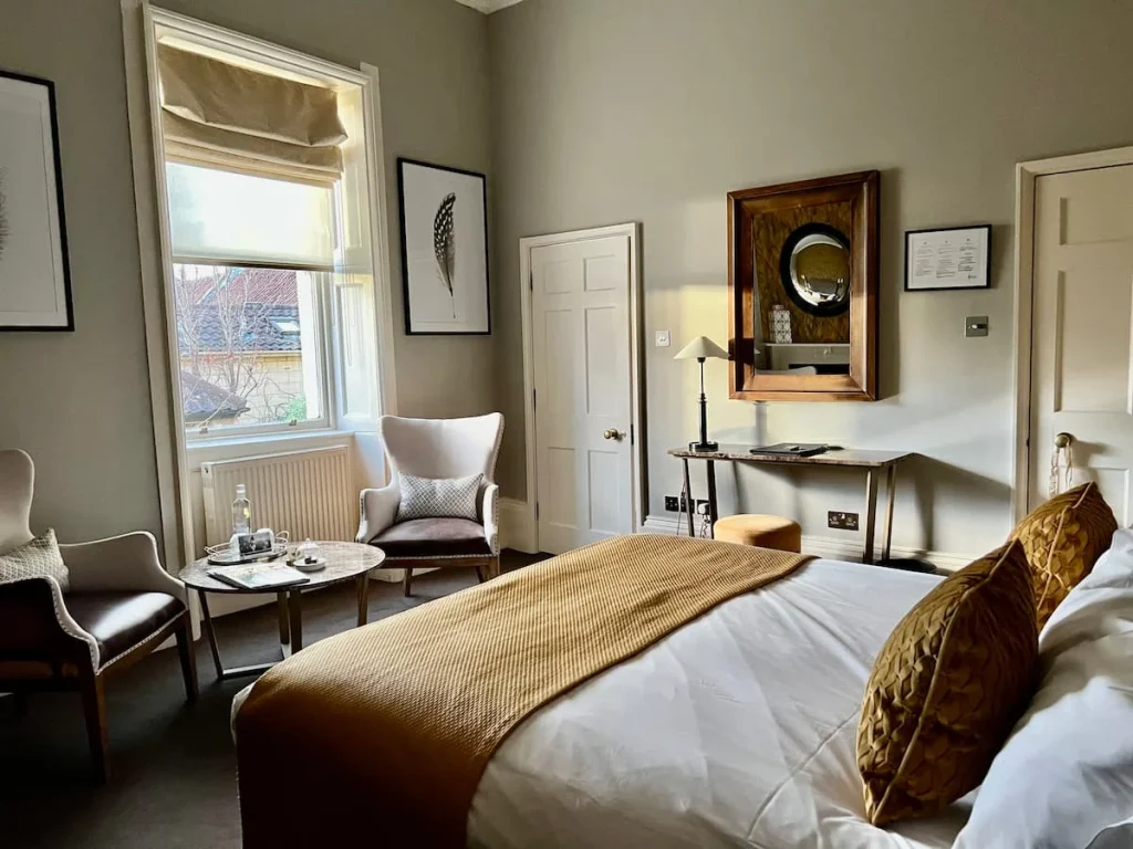 An alternate view of our Superior Room at The Queensberry hotel in Bath. The bed is to the right with a mustard coloured throw and 2 upholstered chairs and a table. The window has matching Roman blinds and there's a small dressing table too