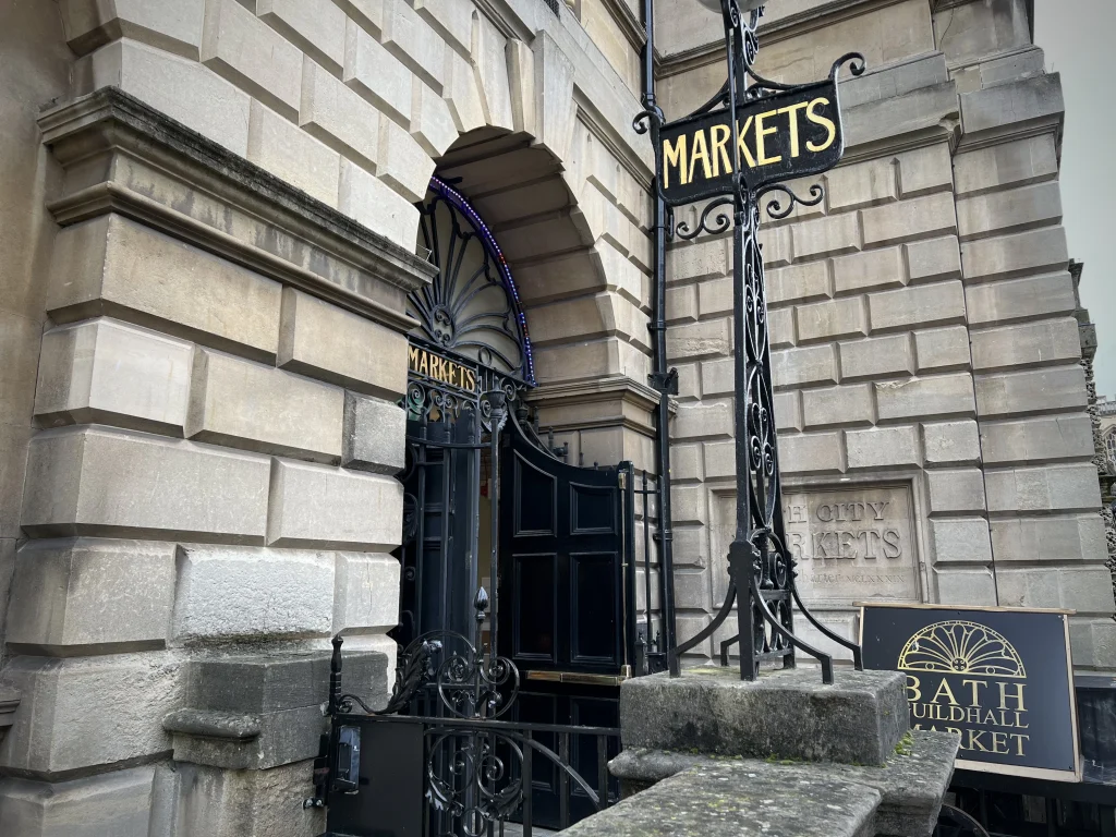 The exterior of The Bath Guildhall Market building which is of a Regency style and has a black sign with gold lettering.