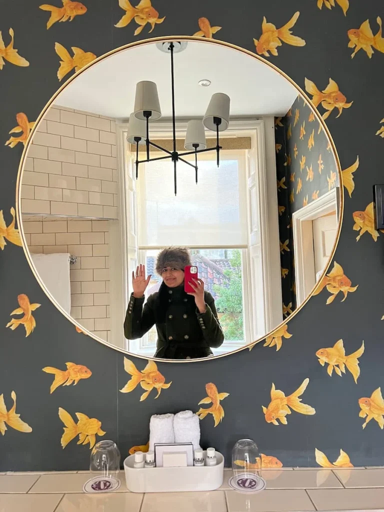 Bejal waving in the bathroom mirror at The Queensberry Hotel, Bath. Bejal's reflection is in a gold rimmed round missor and the wallpaper is dark grey with mustard coloured gold fish.