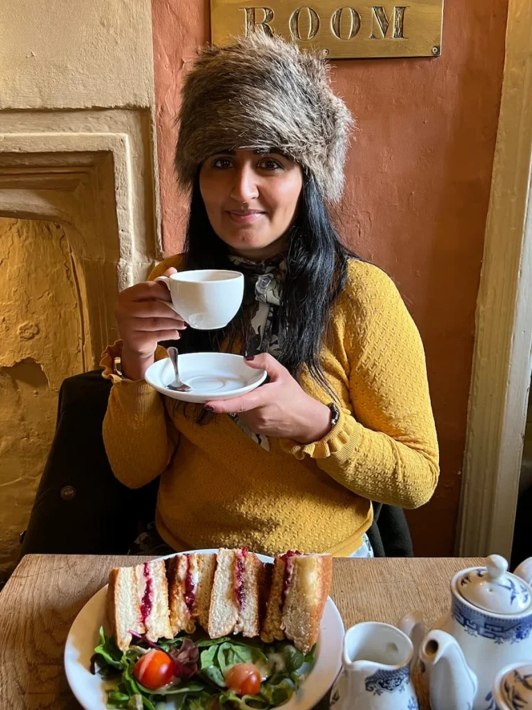 Bejal wearing a Russian style faux fur hat with a mustard jumper holding a teacup and saucer in her hand. on the table is a brie and cranberry bun with salad, at Sally Lunn's Eating House in Bath, England