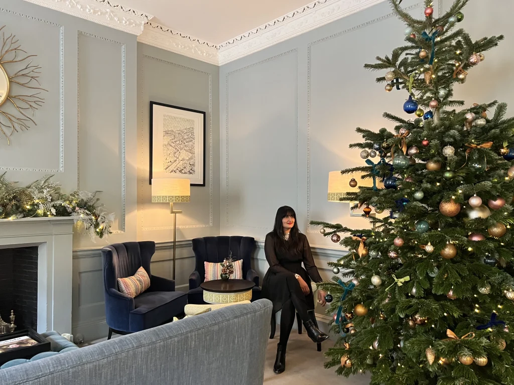 Bejal sitting in front of the Christmas tree wearing a black dress with with lace collars. She is looking at the christmas tree that is decorated with gold, white and blue baubles to match teh same decor in the sitting room at The Queensberry HOtel, Bath