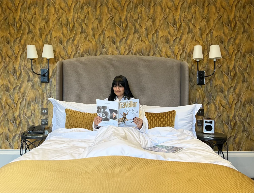 Bejal sitting in bed wearing a white fluffy dressing gown reading Conde Nast Traveller in bed. The bed has white linen and a gold throw at the end. The wallpaper is a mustard and grey wallpaper and the headboard is a light brown colour. There are 2 bedside lights at each of the bed in a superior room at the Queensberry Hotel, a luxury boutique hotel in Bath