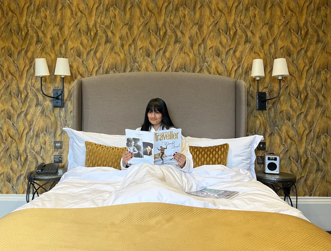Bejal sitting in bed wearing a white fluffy dressing gown reading Conde Nast Traveller in bed. The bed has white linen and a gold throw at the end. The wallpaper is a mustard and grey wallpaper and the headboard is a light brown colour. There are 2 bedside lights at each of the bed in a superior room at the Queensberry Hotel, a luxury boutique hotel in Bath