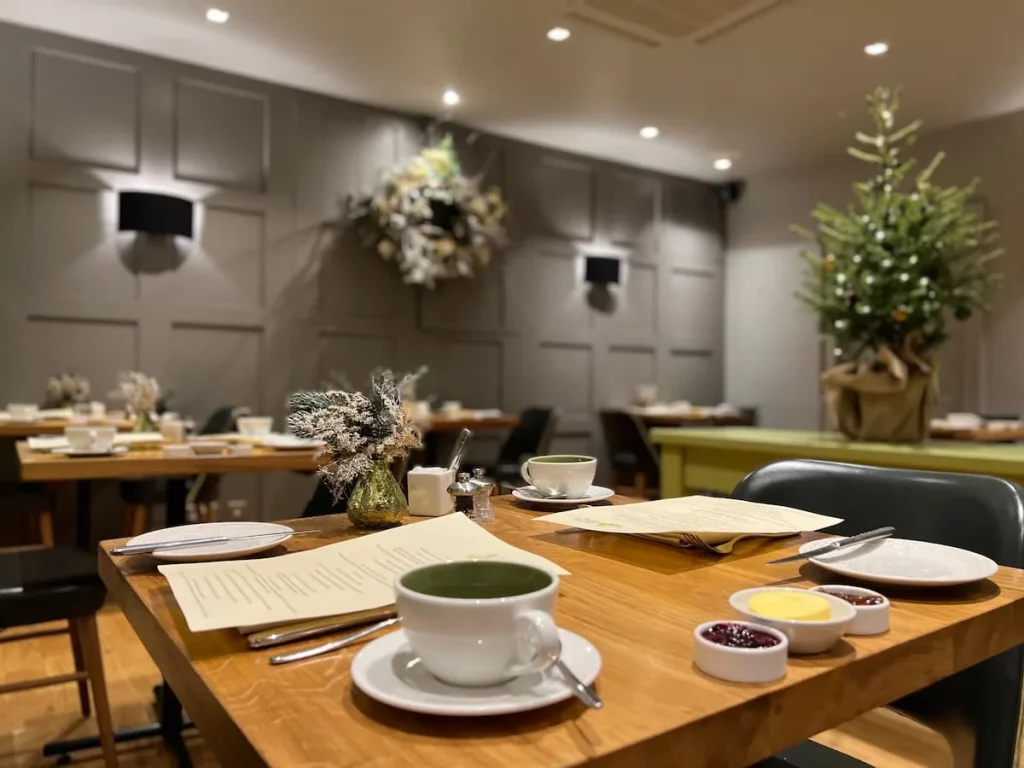 Olive tree restaurant set up for breakfast with Christmas wreathes in the background. There are coffee cups, jams and butter on the tables as well as cutlery and the breakfast menu at The Queensberry Hotel, Bath
