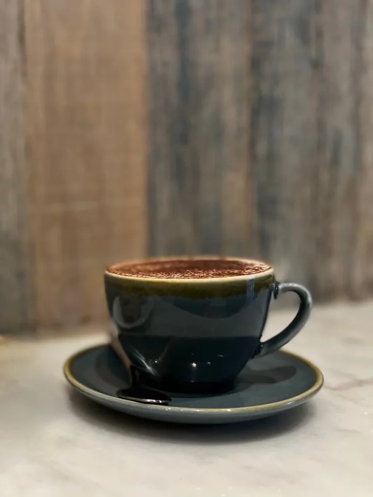 Cappuccino at The Cornish Bakery, Bath. The coffee is in a black coffee cup and saucer against a wood effect background. There are chocolate sprinkles in the cappuccino