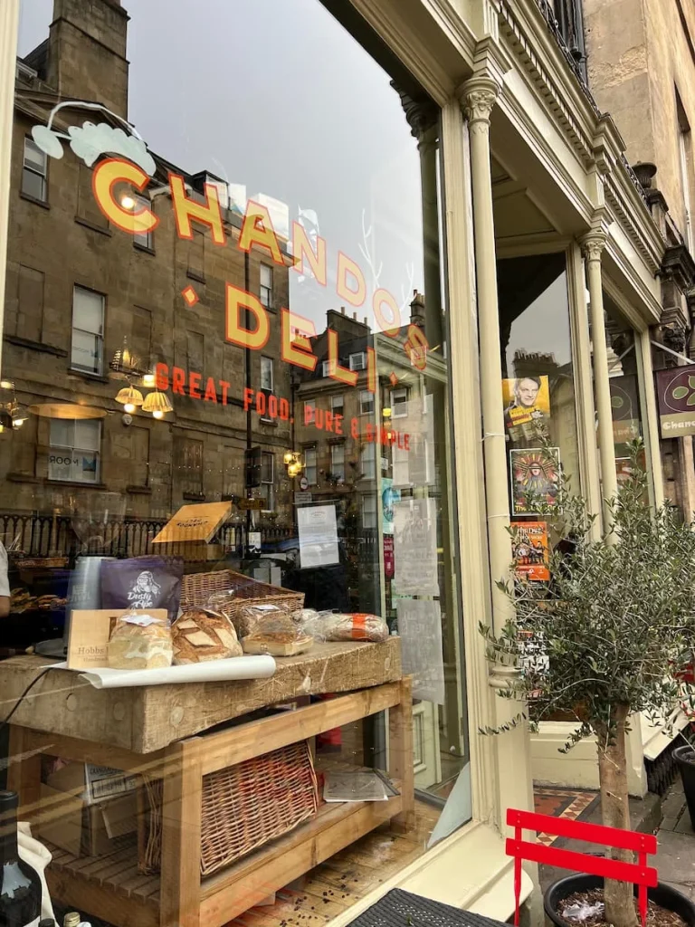 Chandos Exterior in Bath. The writing is in red and there is a green tree planted on the outside. You can also see inside the deli through the large front windows.