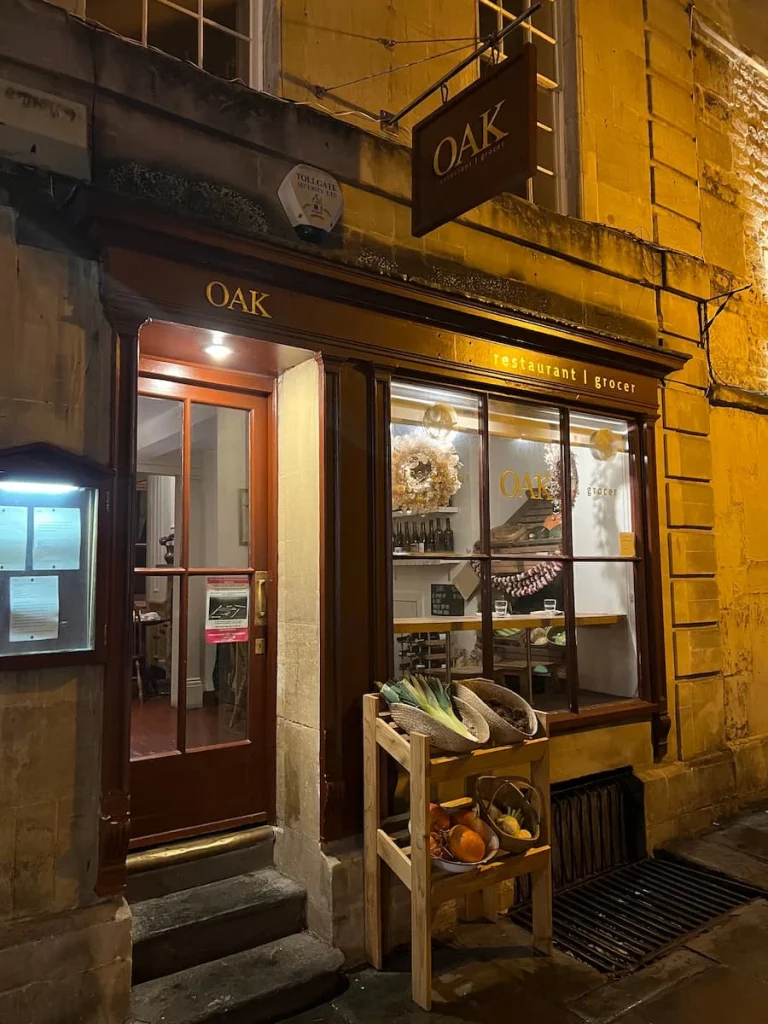 The exterior of AOK restaurant in Bath on an evening. The yellow light from inside reveals an old  shop front with vegetables outside