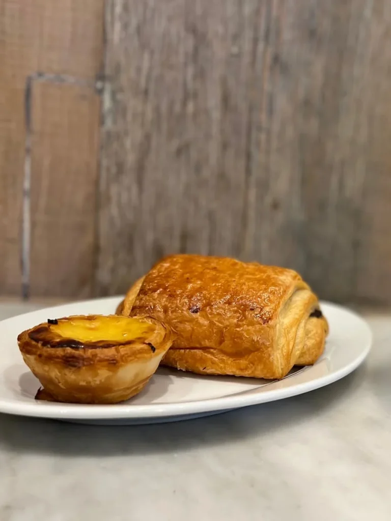 Pain au Chocolat and pastel de nata at The Cornish Bakery, Bath. They are on a white plate against a wood effect background