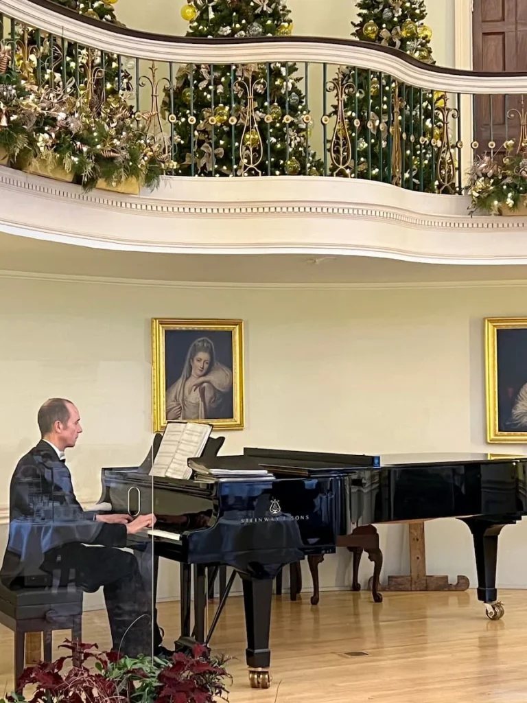 Pianist wearing a black suit playing the grand piano on the stage at the Pump Room Restaurant in Bath. There are festive floral and fairy light displays in the background