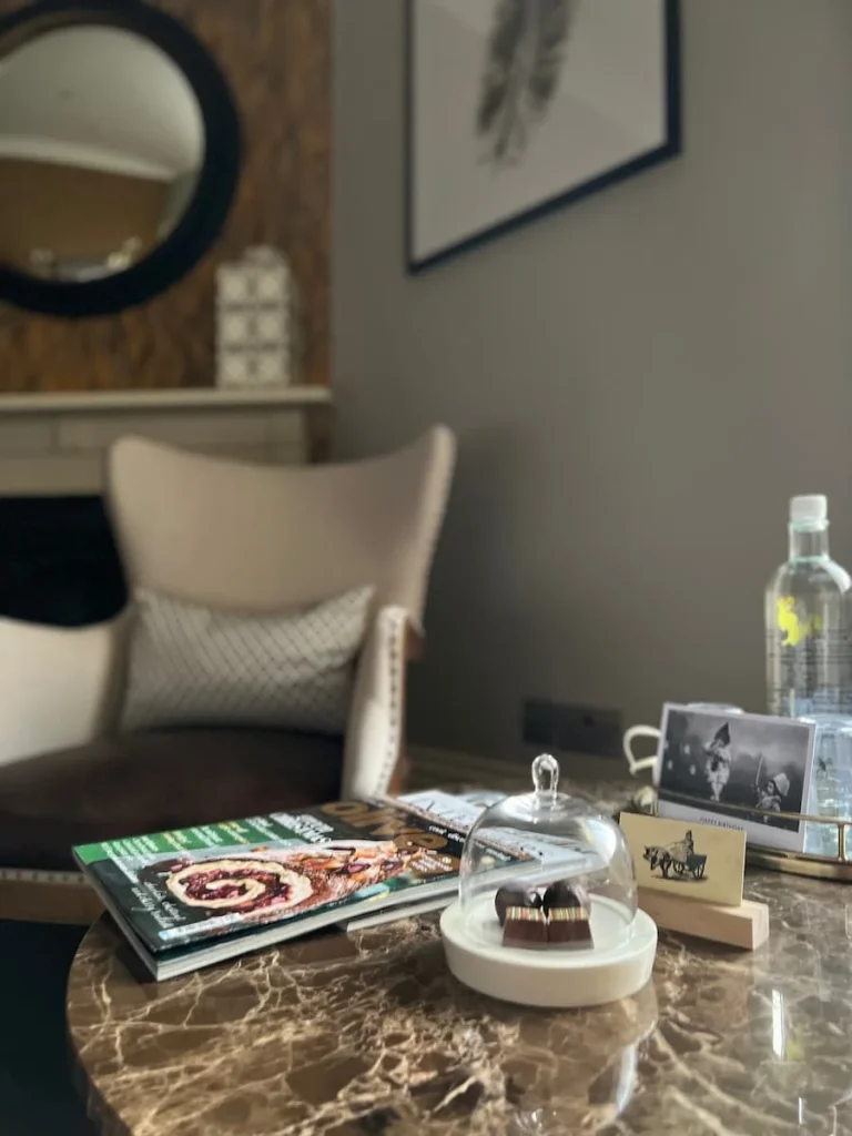 A upholstered chair in the Superior room at The Queensberry Hotel in Bath. There are magazines and truffles on the marble effect table with a water bottle.