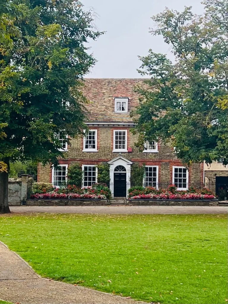 A very cute Dolls house style exterior with a black door, white window frames with intersectional squares and a garden full of multicoloured flowers at the front. There is a black gate around teh house and garden in Ely. A tree frames the image on the right.