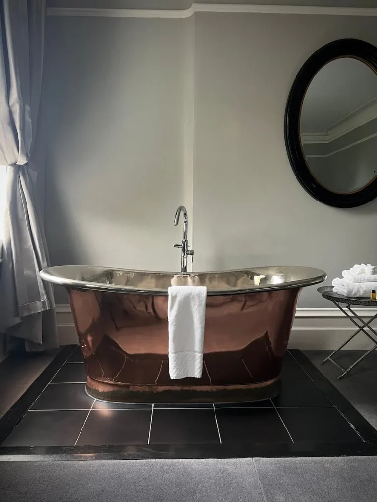 A copper freestanding bath with a white towel draped over the edge at Poet's House Hotel in Ely. There is a oval black framed mirror on the wall and a table with towels and bath products to the side. The window curtains can be seen to the left.