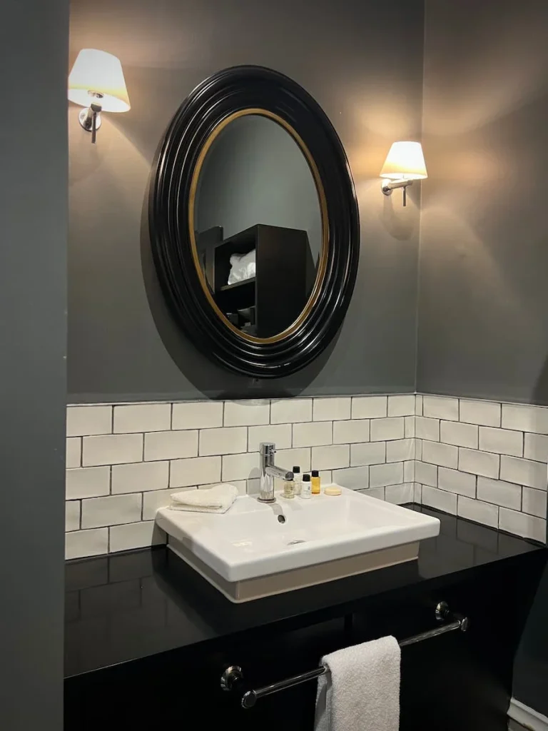 The bathroom vanity area at Poet's House Hotel with a black framed oval mirror on the dark grey wall. The vanity sink is white with toiletteries on it and there are 3 layers of white brick like tiles on the lower part of the wall.