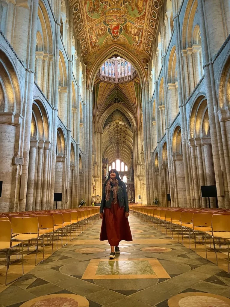 Bejal wearing a burgundy skirt and black leather jacket walking down the Nave of Ely Cathedral. There are rows of chairs at either side and the grand ceiling above with the stone arches to the side.