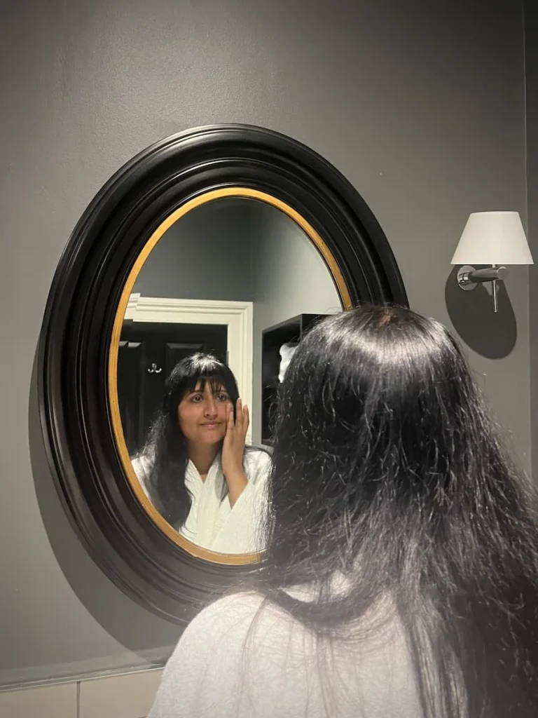 Bejal's reflection in the black framed bathroom mirror at Poet's House luxury boutique hotel in Ely. Bejal is wearing a white fluffy dressing gown and applying lotion to her face. The mirror is on a grey painted wall with a white shaded side lamp