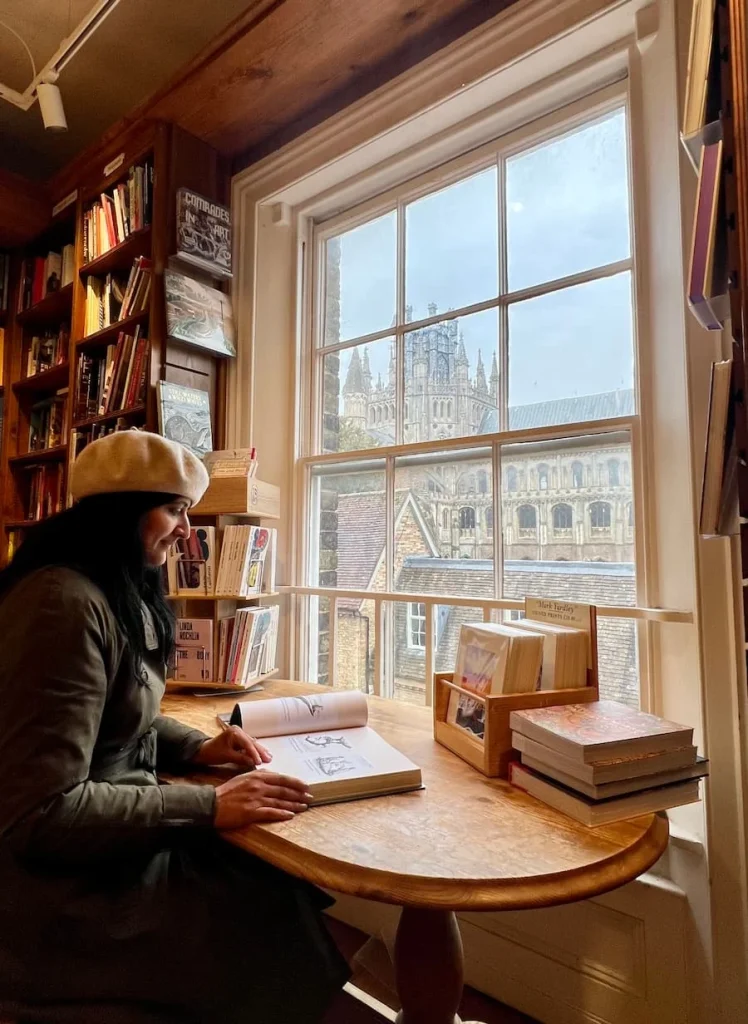 Bejal reading a book at Toppings & Company Bookshop with views out of the Window looking to the Tower of Ely Cathedral. Bejal is wearing a khaki rain mac and a camel coloured beret.