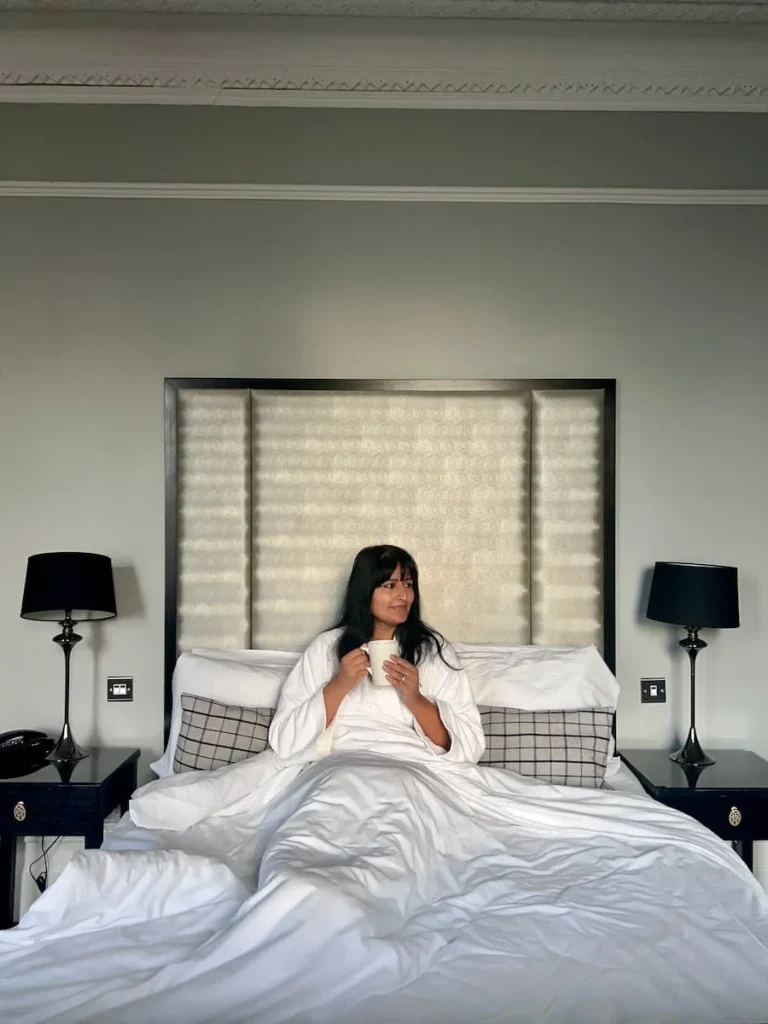 Bejal sitting in bed wearing a fluffy robe with a coffee cup in her gand looking to the left. There are 2 black lamp shaded lights on bedside tables at either side of her and the headboard is tall wuth white and silver framing
