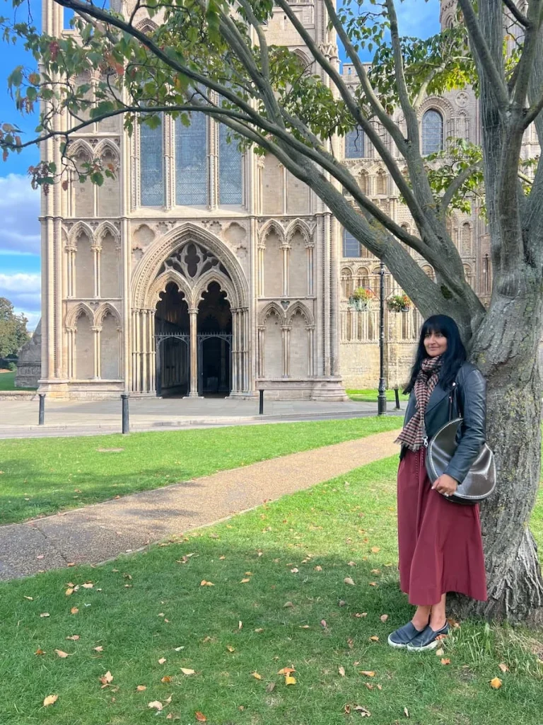 Bejal standing against a tree wearing a burgundy skirt and black leather jacket with black platform pumps. Ely Cathedral is in the background with bright blue skies behind.