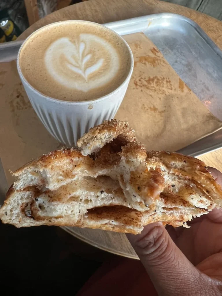 Cappuccino and cardamom bun at The Bake Shop General Store in Ely. The coffee has floral art on it in a white cop and the cardamom bun has a bite taken out of it