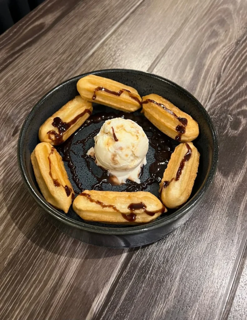 A round dish with 6 churros arranged in a circle, drizzled with chocolate sauce with a scoop of ice-cream in the middle