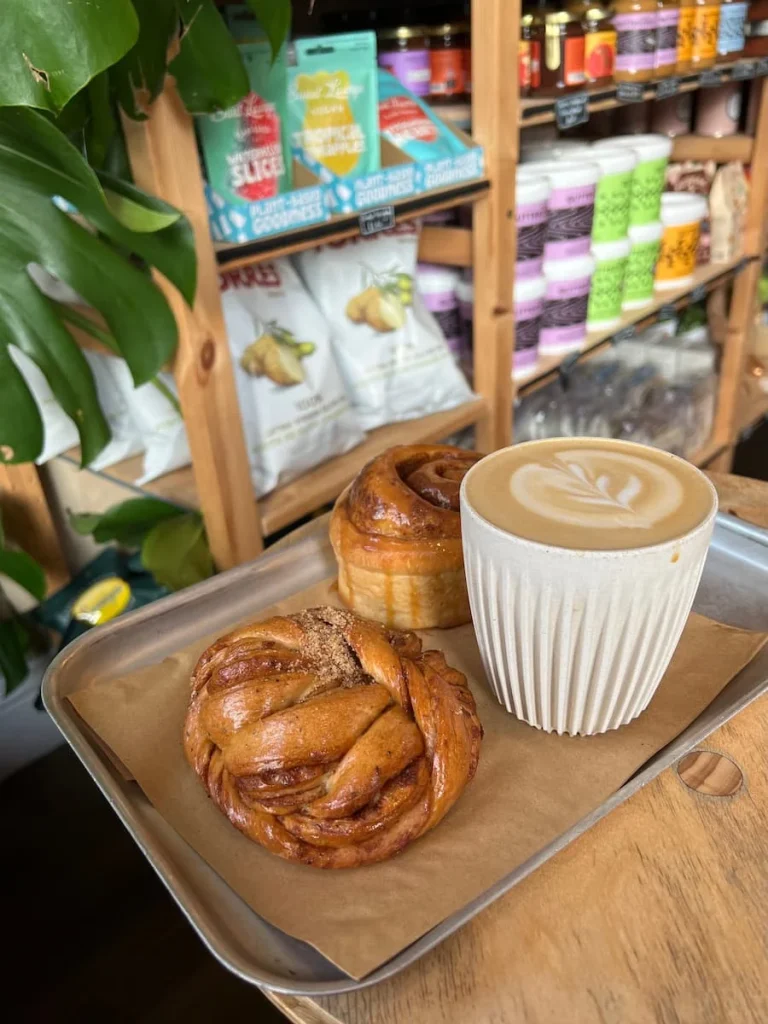 Cinnamon bun, Cardamom bun and cappuccino at at Bake Shop General Store in Ely. They are in a silver tray and in teh background there are packets of food of theshelves