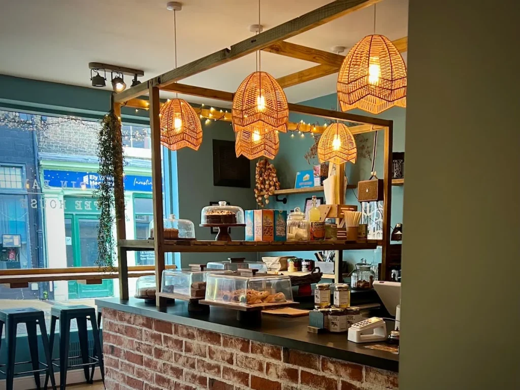 Coffee counter at Tamarin Bay Coffee House, Ely. The counter has various cakes and savoury treats on the counter and light rattan hanging shades with soft yellow light windows.