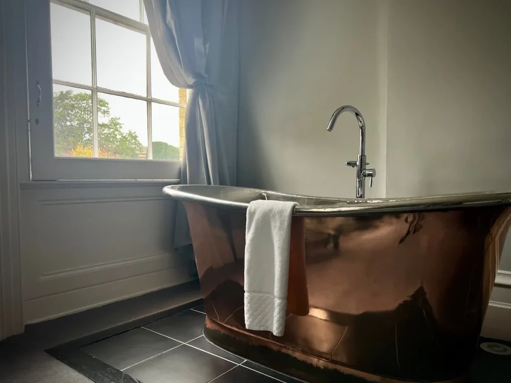 An alternate view of the copper bath with the tap running and window with silk curtain at Poet's house luxury boutique hotel in Ely. Treetops can be seen from the window with the darker morning light coming in. Thee is a white towel mat folded over the bath rim. The walls are a light grey colour.