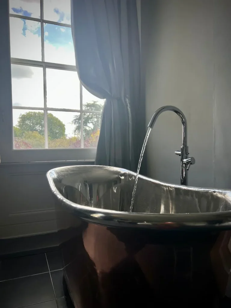 An alternate view of the copper bath with the tap running and window with silk curtain at Poet's house luxury boutique hotel in Ely. Treetops can be seen from the window with the darker morning light coming in. The walls are a light grey colour.