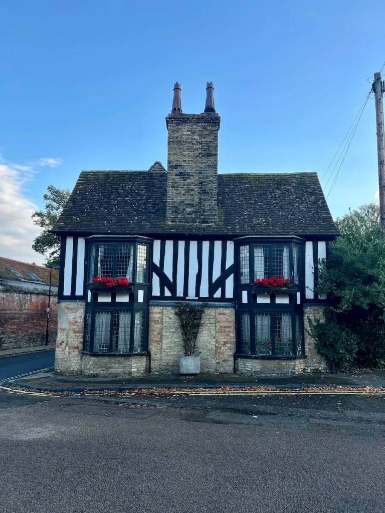 A cute cottage to the side of Ely Cathedral with Tudor black and white timbers and red flower boxes in the windows. There are trees on the side.