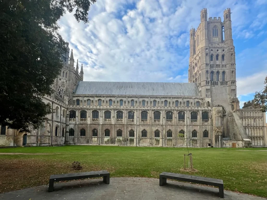 A side view of Ely Cathedral featuring it's full side length. There are 2 brown benches at either side of the image and a freshly mown lawn area to the front of theCathedral