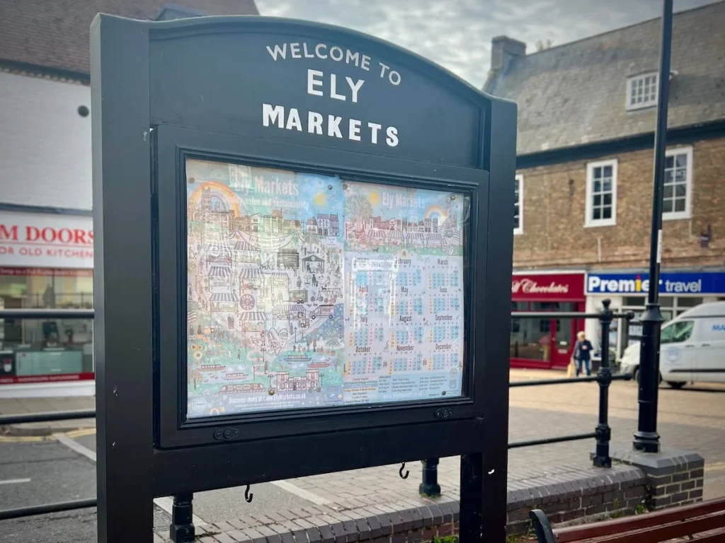 The Ely Markets sign in Market place, Ely. The board is dark grey with a map in teh middle and lettering in white