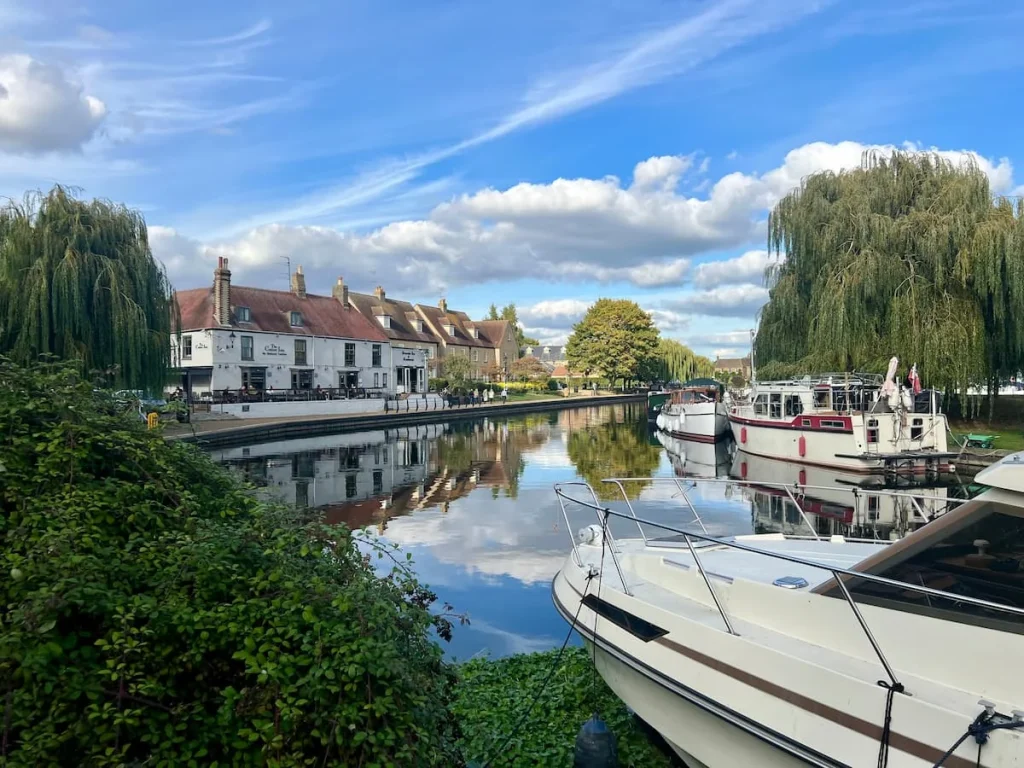 Ely Riverside with barge boats and yachts moored around the edges with bright blue skies and fluffy white cotton wool clouds. The Cutter Inn pub buildings are to the left hand side and there is a reflection of the trees and buildings in the Great River Ouse