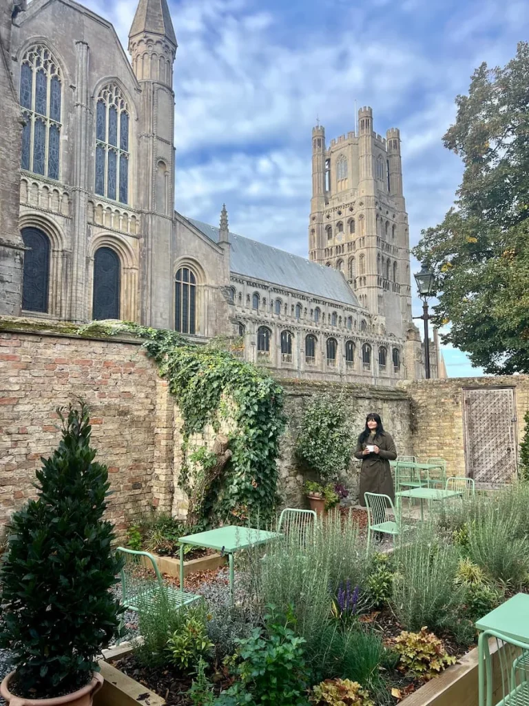 Ely cathedral views from Gail's Bakery garden. Bejal is walking down garden with a cup of coffee in had and the cathedral tower is behind her with foliage in the garden and lavender plants