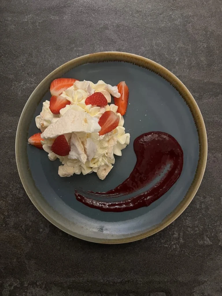 A top lay Deconstructed Eton Mess on a plate with cream, meringue bits and strawberries on a grey plate. There is a swoosh of strawberry coulis on the side.