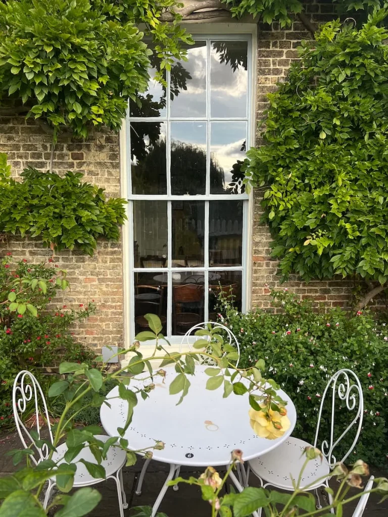 Exterior of Peacocks Tearoom, Ely. A long square paned window with white bistro table and chairs outside with foliage all around