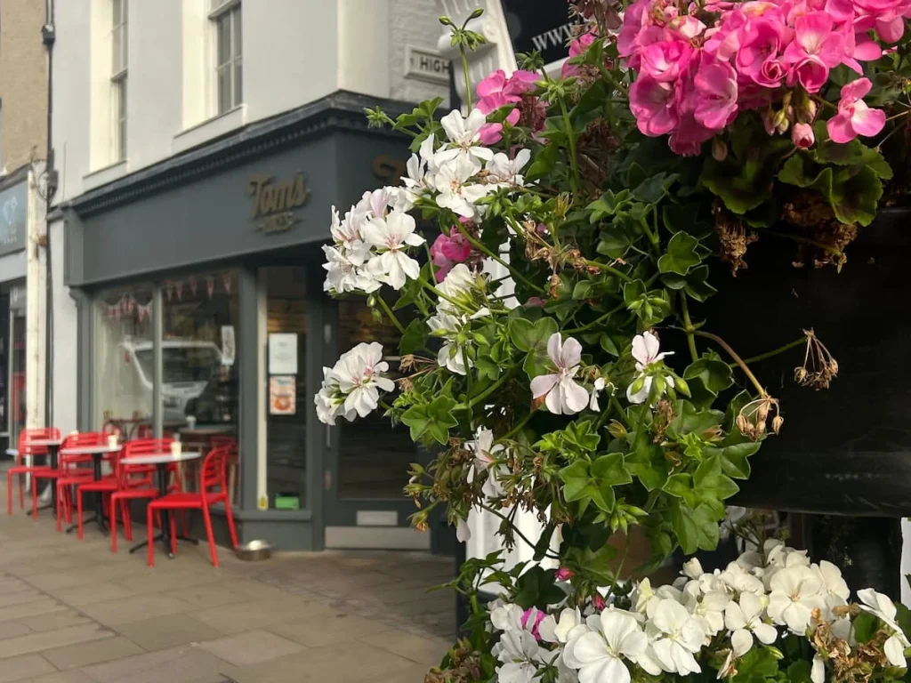 Exterior of Tom's Cafe, Ely. There are pink and white flowers to the right and Tom's cafe grey exterior to the left with red chairs and small bistro tables outside the shop front