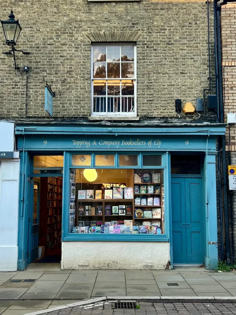 Exterior of Topping & Co, Ely on High Street. The light blue exterior has two doors and the entrance door is open with books in the window that has the lights on as it's a dark rainy day in Ely.