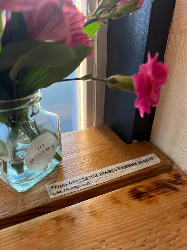 Friends message on wooden counted at Ely Grazing Company. There is a small vase with pink flowers in it and the message is on a strip of white paper and reads, 'True friends are always together in spirit' L.M Montgomery