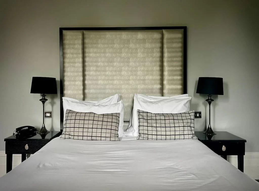 The bed at Poet's House hotel with long cream and silver headboard, Egyptian cotton white bedding and bedside dark wood tables with black lampshades on the lamps at either side of the bed. The walls are a light grey colour