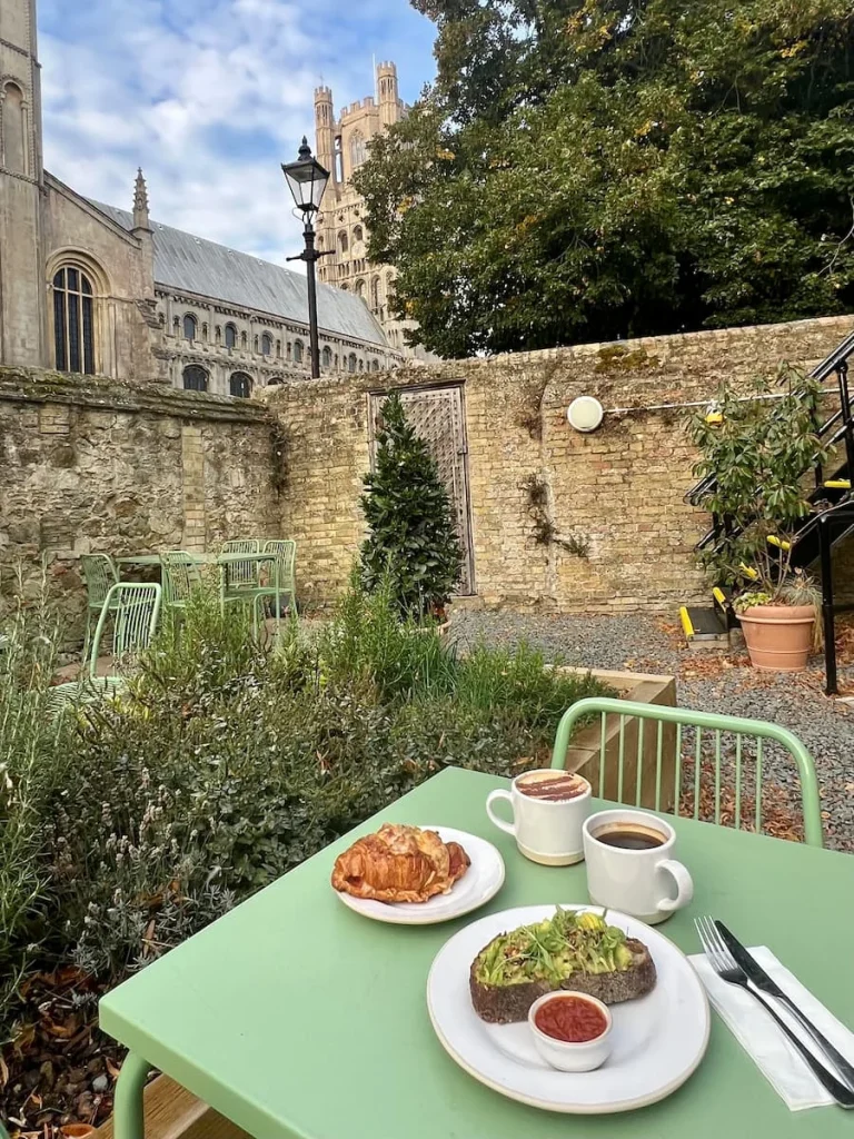 Garden with brunch at Gail's Bakery, Ely. There is avocado bread and a croissant with a cappuccino and black coffee on the light green top table. The background has the tower of Ely Cathedral and the walled garden.