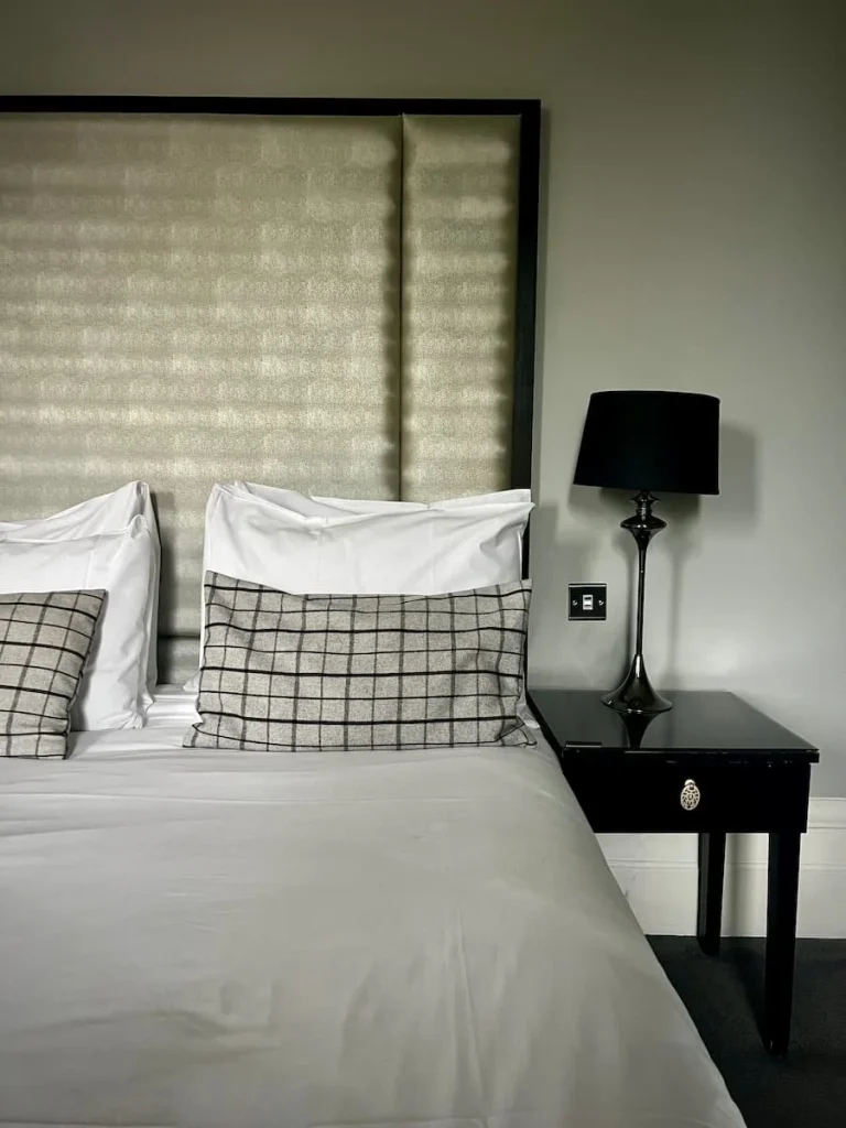 A view of half of the bed at Poet's House hotel with long cream and silver headboard, Egyptian cotton white bedding and bedside dark wood table with black lampshade on the lamp