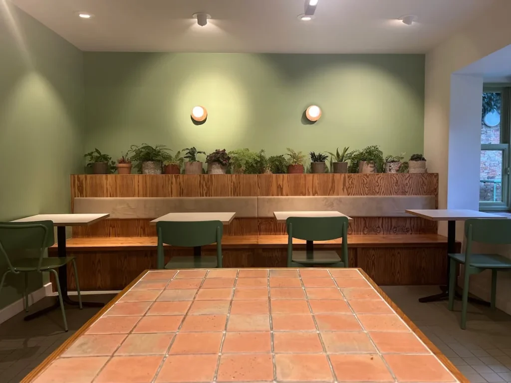 Indoor seating at Gail's bakery in Ely. The walls are a pale green with small plants in pots along the back wall. The tables area. light wood with green chairs tucked under the three tables