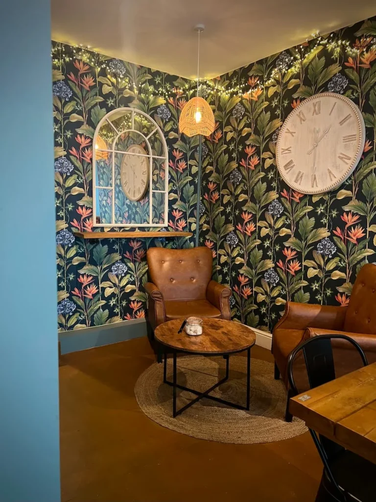 Interiors of Tamarin Bay Coffee House in Ely. There is a floral teal , pink and yellow wallpaper on the walls with two brown leather comfy seats with a small table between each. A large wall clock hangs on the wallpaper as well as a big window style white mirror.