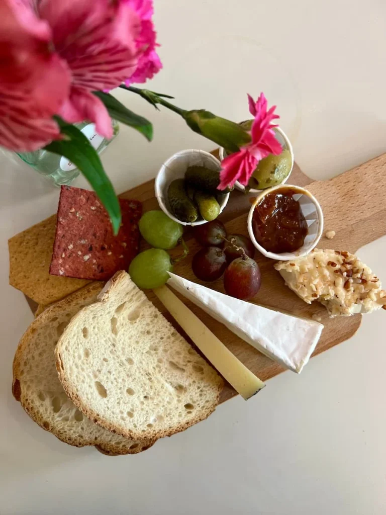 A flat lay of the mini 3 cheese board with slices of bread, grapes, three cheeses, gherkins chutney and crackers on a brown wooden board. There are pink flowers in a small vase on the side.