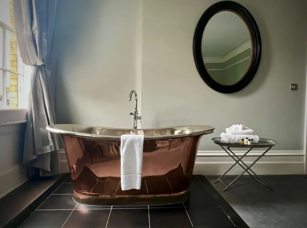 A copper freestanding bath with a white towel draped over the edge at Poet's House Luxury Boutique Hotel in Ely. There is a oval black framed mirror on the wall and a table with towels and bath products to the side. The window curtains can be seen to the left.