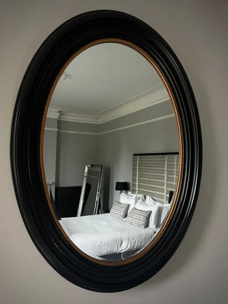 A dark brown rimmed oval mirror with the reflection of the bed, free standing mirror and wardrobe at Poet's House House luxury boutique hotel in Ely
