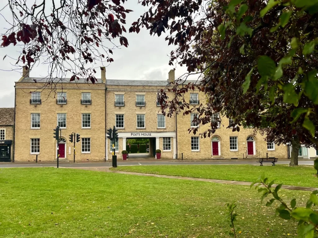 The pale sand coloured exterior of the historic building that is now the Poet's House Luxury Boutique  Hotel in Ely. There is a crossing outside the hotel and a public green lawn. There is a small courtyard in the centre with windows and red doors to either side