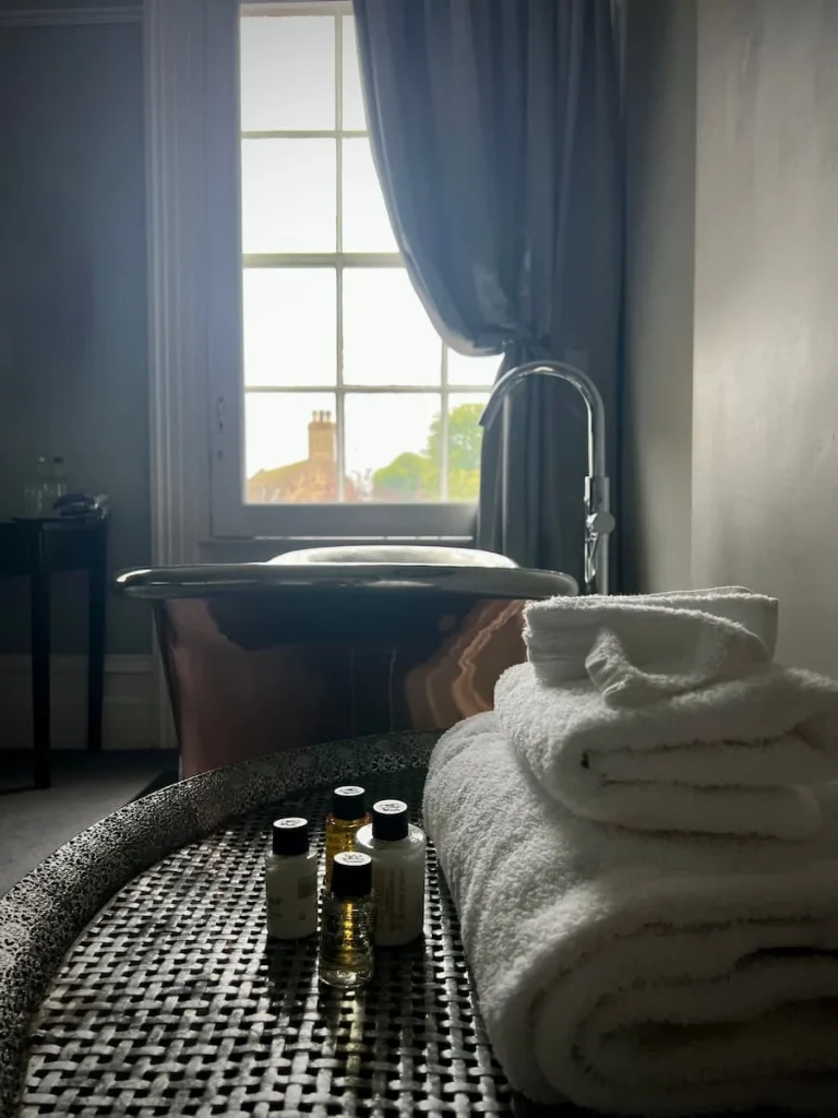 An alternative view of the copper bath from the side looking towards the window. there are a pile of towels and bath products on the small table. The low evening light is shining into the room, causing a slightly dreamy low light effect